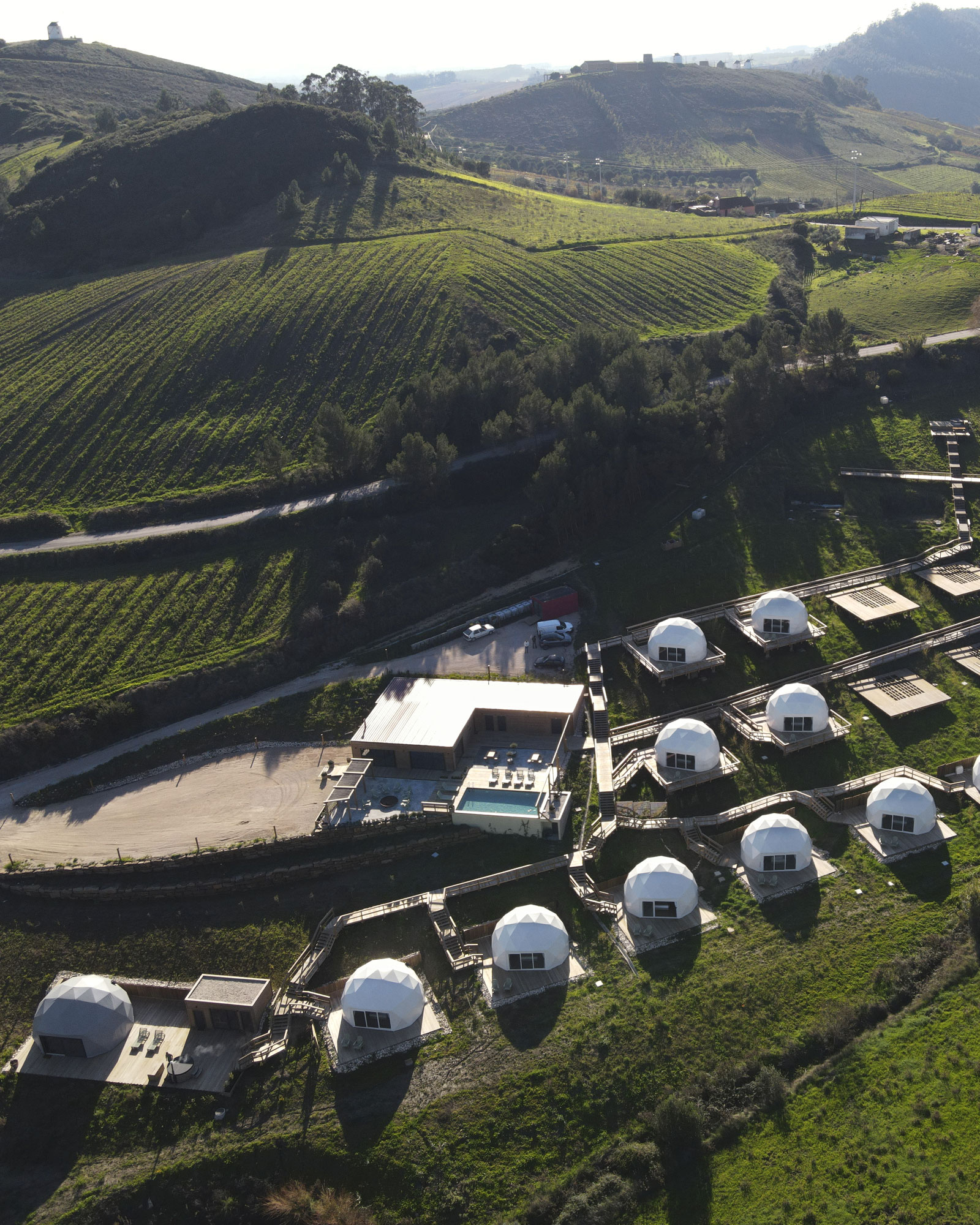 Aerial view of a hillside resort with white geodesic dome cabins connected by wooden walkways and a central building with a pool, surrounded by green fields and hills.